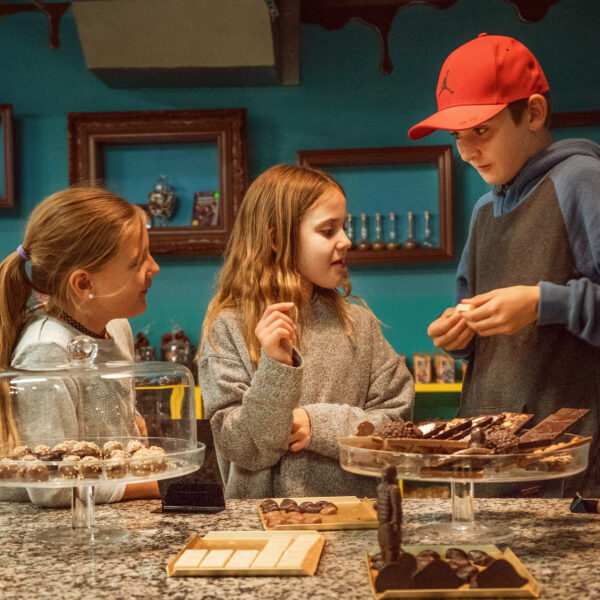 Drei Kinder stehen an der Theke eines Schokoladenladens, unterhalten sich und wählen Pralinen aus verschiedenen Auslagen aus.