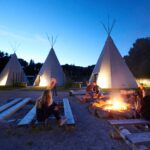 Menschen sitzen in der Abenddämmerung auf Holzbänken um ein Lagerfeuer herum, im Hintergrund stehen mehrere große Tipis, die von Lichtern beleuchtet werden.