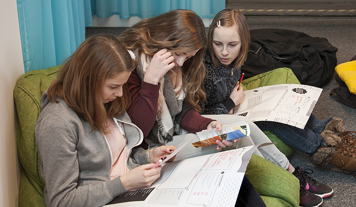 Drei Mädchen im Teenageralter sitzen auf dem Boden, sehen sich Papiere und Zeitschriften an und scheinen gemeinsam an einem Projekt oder einer Aufgabe zu arbeiten.