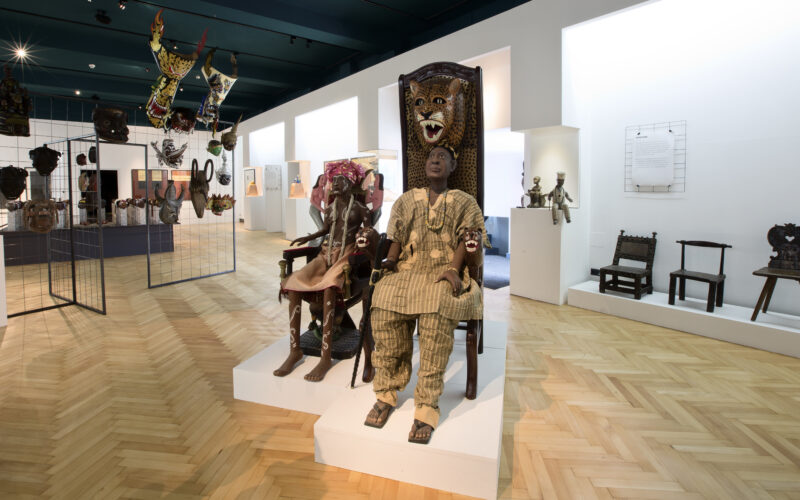 Eine Museumsausstellung zeigt afrikanische Stühle, Masken und Skulpturen, darunter zwei Schaufensterpuppen in traditioneller Kleidung, die auf kunstvollen Thronen sitzen.