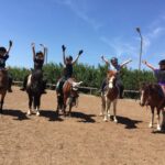 Fünf Kinder mit Helmen sitzen in einer Reihe auf Pferden und heben ihre Arme in die Luft, auf einem unbefestigten Reitplatz mit Bäumen und einem blauen Himmel im Hintergrund.