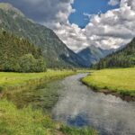 Ein klarer Fluss fließt durch ein grünes Tal, das von dichten Wäldern und hohen Bergen unter einem teilweise bewölkten Himmel umgeben ist.
