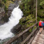 Zwei Wanderer mit Rucksäcken überqueren eine Holzbrücke neben einem rauschenden Wasserfall in einer bewaldeten, felsigen Landschaft.