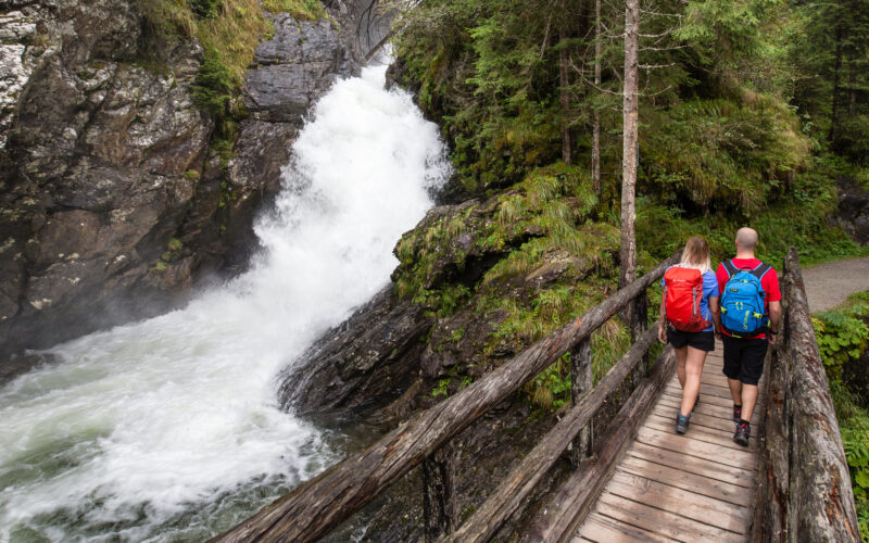 Zwei Wanderer mit Rucksäcken überqueren eine Holzbrücke neben einem rauschenden Wasserfall in einer bewaldeten, felsigen Landschaft.