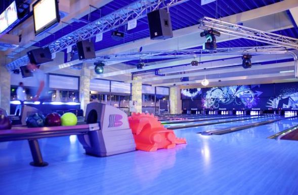 Modernes Interieur einer Bowlingbahn mit mehreren Bahnen, bunten Bowlingkugeln auf einem Gestell, orangefarbenen Kunststofframpen und stimmungsvoller Beleuchtung.