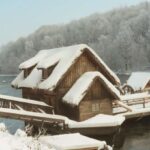 Eine schneebedeckte hölzerne Wassermühle steht an einem Fluss mit einem schneebedeckten Wald im Hintergrund.