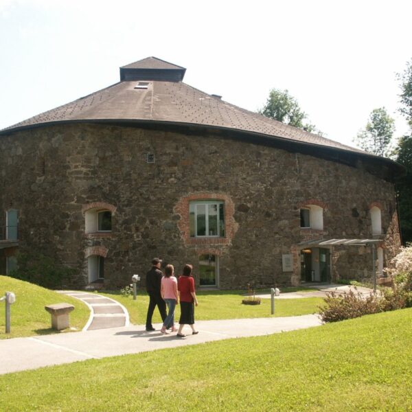 Drei Personen gehen auf einem Weg zu einem großen, runden Steingebäude mit kleinen Fenstern und einem schrägen Dach, umgeben von Gras und Bäumen.