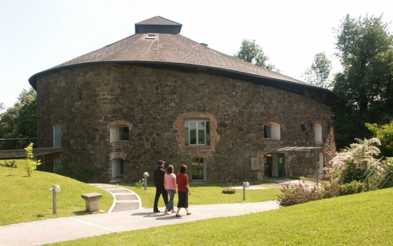 Drei Personen gehen auf einem Weg zu einem großen, runden Steingebäude mit kleinen Fenstern und einem schrägen Dach, umgeben von Gras und Bäumen.