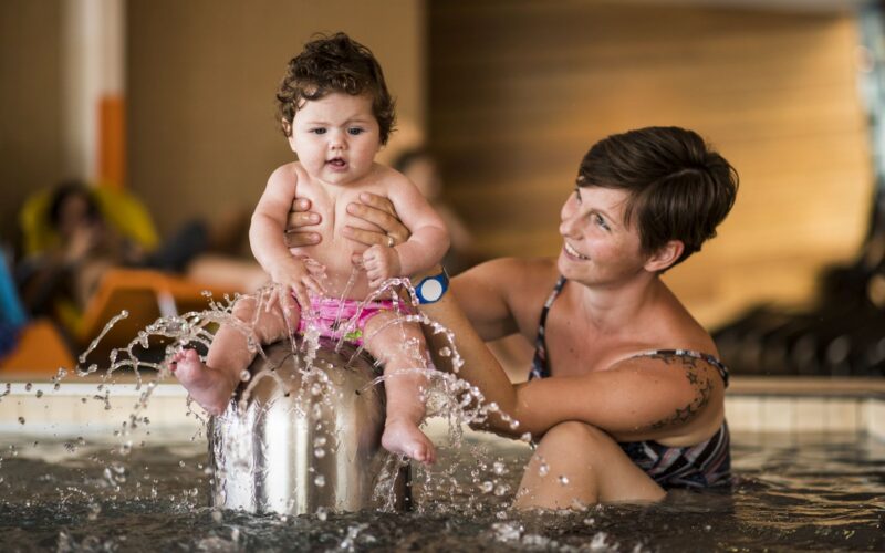 Ein Erwachsener hält ein Baby, das auf einem Springbrunnen in einem Schwimmbecken sitzt, wobei das Wasser plätschert und beide die Aktivität zu genießen scheinen.