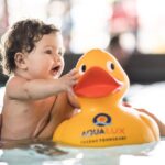 Ein Baby in einem Schwimmbecken greift nach einer großen gelben Gummiente mit der Aufschrift "AQUALUX", während es von einer Erwachsenenhand gestützt wird.