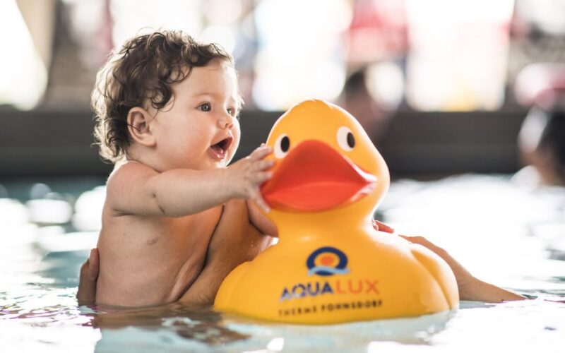 Ein Baby in einem Schwimmbecken greift nach einer großen gelben Gummiente mit der Aufschrift "AQUALUX", während es von einer Erwachsenenhand gestützt wird.