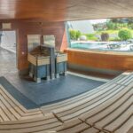 Weitwinkelansicht einer leeren Holzsauna mit abgestuften Sitzgelegenheiten, einem Zentralofen und einem großen Fenster mit Blick auf einen Außenpool und Liegestühle.