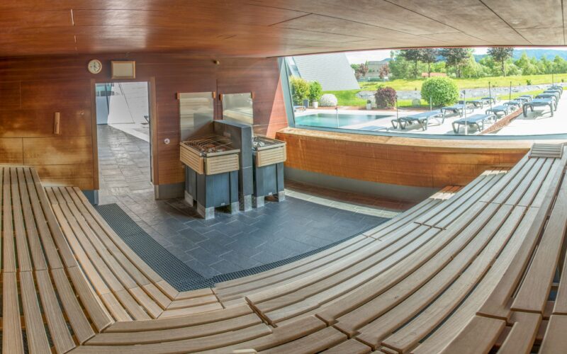 Weitwinkelansicht einer leeren Holzsauna mit abgestuften Sitzgelegenheiten, einem Zentralofen und einem großen Fenster mit Blick auf einen Außenpool und Liegestühle.