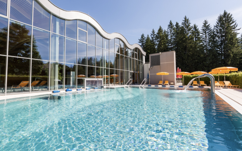 Außenschwimmbad mit klarem Wasser, gelben Liegestühlen und Sonnenschirmen neben einem modernen Glasgebäude, umgeben von Bäumen und unter freiem Himmel.