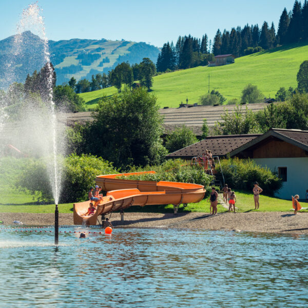 Menschen genießen einen sonnigen Tag am Strand eines Sees mit einer Wasserrutsche, einem Springbrunnen und grasbewachsenen Hügeln im Hintergrund.