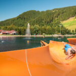 Ein Kind rutscht eine orangefarbene Wasserrutsche hinunter in einen natürlichen Badesee, mit schwimmenden Menschen und einer bewaldeten Berglandschaft im Hintergrund.