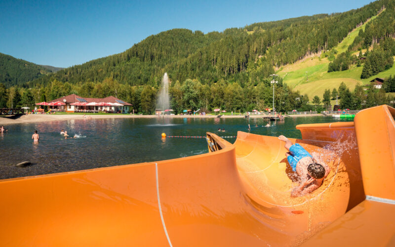 Ein Kind rutscht eine orangefarbene Wasserrutsche hinunter in einen natürlichen Badesee, mit schwimmenden Menschen und einer bewaldeten Berglandschaft im Hintergrund.