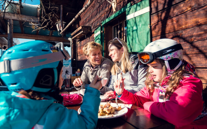 Zwei Erwachsene und zwei Kinder in Skikleidung sitzen an einem Holztisch im Freien und genießen das Essen an einem sonnigen Tag neben einem rustikalen Holzgebäude.