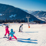 Zwei Kinder in Skiausrüstung üben auf einer verschneiten Piste in der Nähe einer pinguinförmigen Markierung das Skifahren, mit Bergen und Skiliften im Hintergrund.