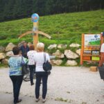 Eine Gruppe von Menschen steht im Freien und hört einem Sprecher in der Nähe eines Informationsdisplays und einer Holzkonstruktion zu, mit einem grasbewachsenen Hügel und Felsen im Hintergrund.