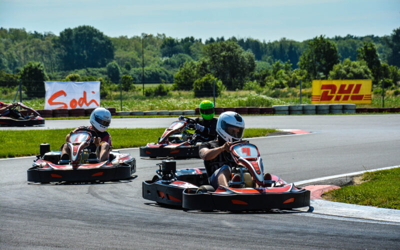 Drei Personen mit Helmen fahren Go-Karts um eine Kurve auf einer Freiluftstrecke, im Hintergrund sind Bäume und Werbebanner zu sehen.