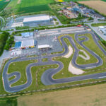 Luftaufnahme einer Go-Kart-Rennstrecke mit gewundenen Asphaltbahnen, umgeben von Gras, Gebäuden, Parkplätzen und Ackerland.