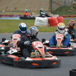 Vier Personen in Rennanzügen und mit Helmen fahren mit Gokarts um eine Kurve, während im Hintergrund zwei weitere Fahrer auf einer Rennstrecke im Freien zu sehen sind.