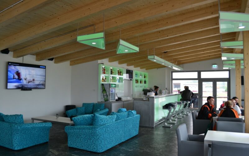 Moderne Lounge mit tealfarbenen Sofas, einem an der Wand montierten Fernseher, einem Barbereich mit Hockern und einigen Tischen unter einer Holzbalkendecke.