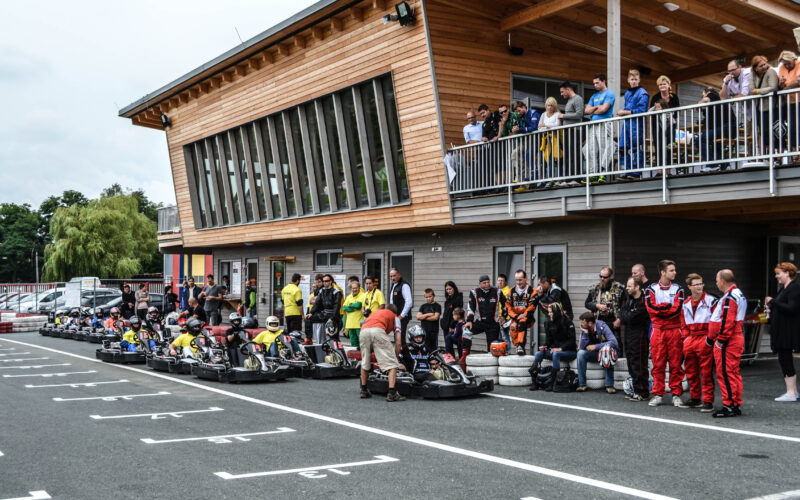 Die Menschen versammeln sich und sehen zu, wie sich die Go-Kart-Fahrer in der Startaufstellung vor einem modernen Holzgebäude mit einem Balkon aufstellen.