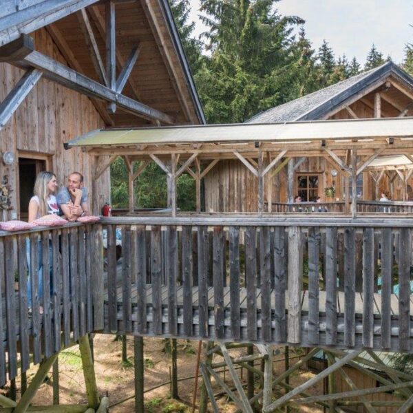 Zwei Personen stehen auf dem Balkon einer Holzhütte, die durch einen überdachten Gang verbunden und von Bäumen umgeben ist.