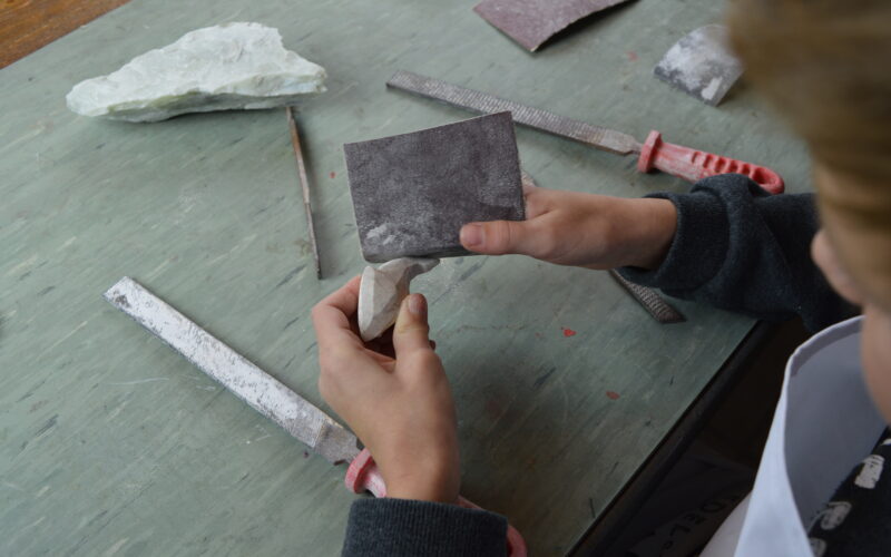 Eine Person glättet mit Schleifpapier und Feilen ein Stück Stein auf einem Tisch, der mit Werkzeugen und einem Steinfragment vollgestopft ist.