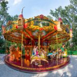 Ein farbenfrohes Karussell mit Kindern und Erwachsenen, die auf verschiedenen Tierfiguren reiten, in einer von Bäumen und Grün umgebenen Parklandschaft.