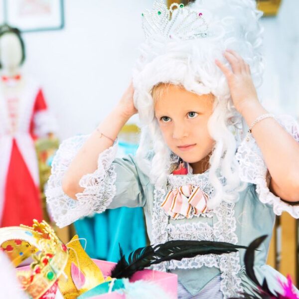 Ein kleines Kind mit weißer Perücke, Krone und historischem Kostüm richtet seine Perücke, umgeben von farbenfrohen Kostümaccessoires.