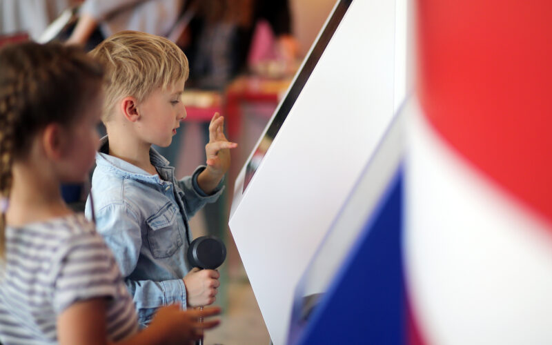 Zwei Kinder interagieren mit einem Touchscreen-Kiosk; der Junge berührt den Bildschirm und hält einen telefonähnlichen Hörer in der Hand, während das Mädchen neben ihm steht.