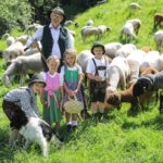 Ein Mann und vier Kinder in traditioneller Kleidung stehen auf einem grasbewachsenen Feld mit einer Schafherde und einem schwarz-weißen Hund.
