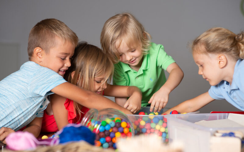 Vier kleine Kinder sind um einen Tisch versammelt und greifen in einen Behälter mit bunten Perlen oder Bonbons, wobei sie engagiert und konzentriert wirken.
