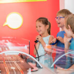 Drei kleine Kinder stehen in einem hell erleuchteten Raum mit roten Wänden um eine Slotcar-Rennbahn herum, lächeln und halten Controller in der Hand.