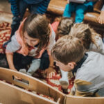 Mehrere Kinder sitzen auf einem gemusterten Teppich, dicht gedrängt um einen hölzernen Gegenstand herum und scheinen konzentriert einer Aktivität im Haus nachzugehen.