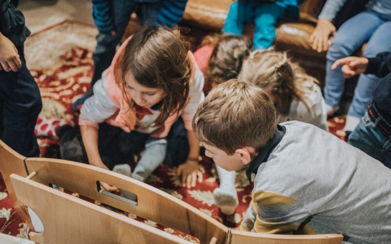 Mehrere Kinder sitzen auf einem gemusterten Teppich, dicht gedrängt um einen hölzernen Gegenstand herum und scheinen konzentriert einer Aktivität im Haus nachzugehen.