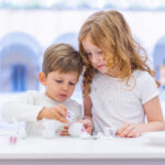 Zwei Kinder sitzen an einem Tisch und feiern eine Teeparty mit einem kleinen weißen und geblümten Spielzeug-Teeservice.