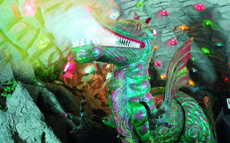 Ein großer animatronischer Drache mit leuchtenden Augen und Maul sitzt in einem Höhlentunnel, der mit bunten Lichtern dekoriert ist.