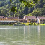 Eine Reihe gelber Gebäude, wahrscheinlich ein Hotel oder eine Ferienanlage, liegt am grünen Ufer eines ruhigen Sees mit einem bewaldeten Hügel im Hintergrund.