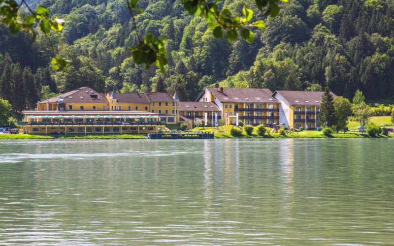 Eine Reihe gelber Gebäude, wahrscheinlich ein Hotel oder eine Ferienanlage, liegt am grünen Ufer eines ruhigen Sees mit einem bewaldeten Hügel im Hintergrund.