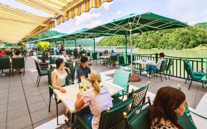Menschen speisen im Freien unter grünen Sonnenschirmen auf einer Terrasse am Flussufer mit Blick auf die Bäume und Hügel im Hintergrund.
