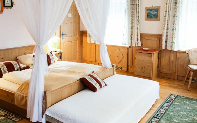 Ein gemütliches Hotelzimmer mit einem Doppelbett und einem Einzelbett, Holzmöbeln, dekorativen Teppichen, Vorhängen und natürlichem Licht durch große Fenster.