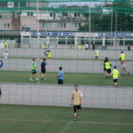 Mehrere Gruppen von Menschen spielen Fußball auf einem Kunstrasenplatz im Freien, getrennt durch Netze, mit Gebäuden im Hintergrund.