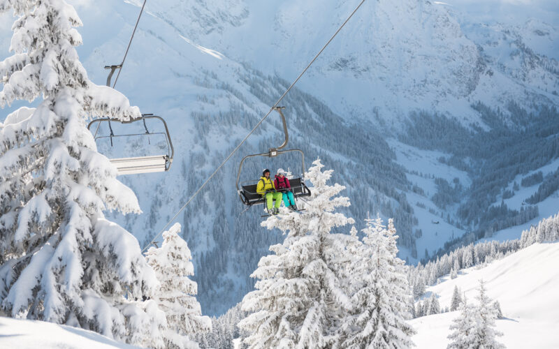 Zwei Personen in Skiausrüstung sitzen auf einem Skilift, der über schneebedeckte Bäume mit Bergen im Hintergrund hinauffährt.