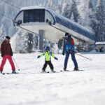 Drei Personen, darunter ein Kind, fahren auf einem verschneiten Hang in der Nähe einer Skiliftstation Ski, im Hintergrund sind schneebedeckte Bäume zu sehen.