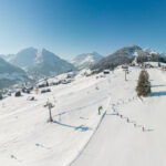 Luftaufnahme einer verschneiten Skipiste mit Skifahrern, Skilift und verstreuten Gebäuden; umgeben von Bergen und Kiefern unter einem klaren blauen Himmel.