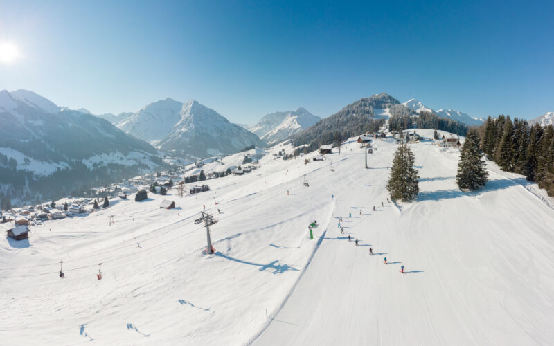 Luftaufnahme einer verschneiten Skipiste mit Skifahrern, Skilift und verstreuten Gebäuden; umgeben von Bergen und Kiefern unter einem klaren blauen Himmel.